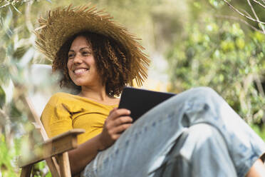 Smiling woman in straw hat holding digital tablet while sitting on chair in vegetable garden - SBOF03095