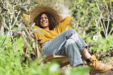 Smiling woman in straw hat with hands behind head relaxing on deck chair in garden - SBOF03096
