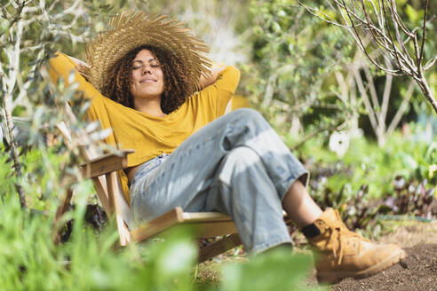 Smiling woman in straw hat resting on chair in garden - SBOF03097