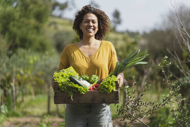 Smiling woman holding crate of fresh vegetables while standing in garden - SBOF03102