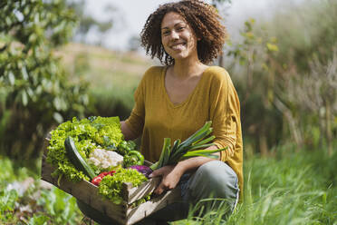 Smiling woman squatting while holding crate of fresh vegetables in garden - SBOF03103