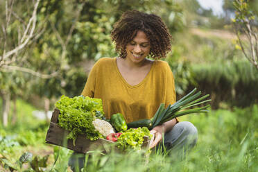 Lächelnde Frau betrachtet Gemüse in einer Kiste, während sie im Garten hockt - SBOF03104