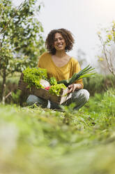 Smiling woman squatting while holding leafy vegetables against sky in garden - SBOF03106