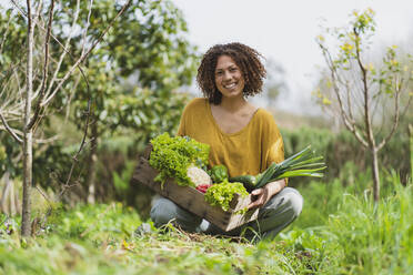 Smiling curly haired woman holding wooden crate of vegetables while squatting in garden - SBOF03107
