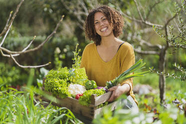 Smiling woman holding a crate with freshly picked organic vegetables in permaculture garden - SBOF03111