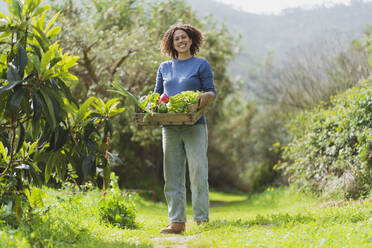 Smiling woman with vegetables crate standing in garden - SBOF03118