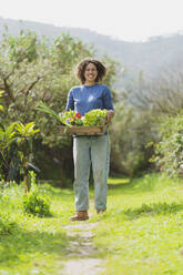 Smiling woman with crate of fresh green vegetables standing in garden - SBOF03119