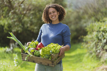 Smiling woman holding crate of vegetables in permaculture garden - SBOF03120