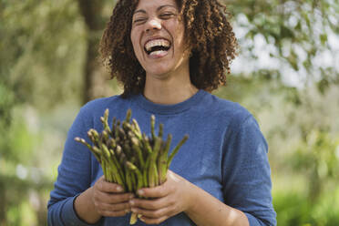 Laughing woman holding fresh green asparagus in permaculture garden - SBOF03122