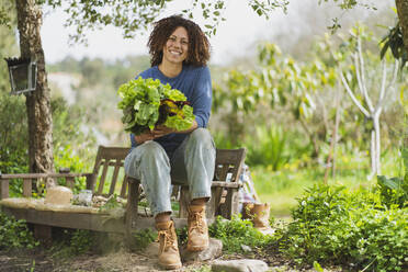 Smiling woman holding leafy vegetables while sitting on wooden bench in garden - SBOF03123