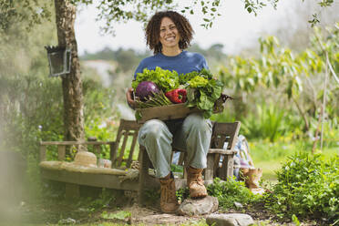 Lächelnde Frau, die eine Kiste mit frischem grünem Gemüse hält, während sie auf einer Bank im Garten sitzt - SBOF03124