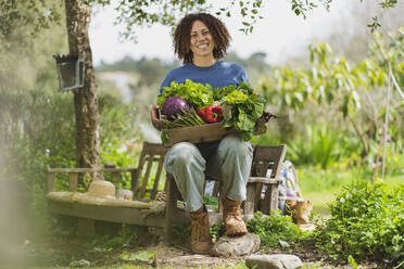 Smiling woman holding crate with fresh green vegetables while sitting on bench in garden - SBOF03124