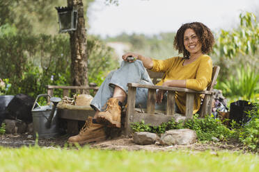 Lächelnde Frau hält eine Schere und sitzt auf einer Holzbank im Garten - SBOF03126