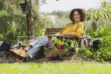 Junge lächelnde Frau sitzt auf einer Holzbank in einem nachhaltigen Permakultur-Garten - SBOF03127