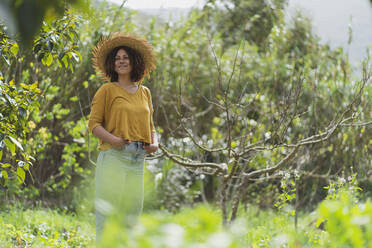 Lächelnde Frau mit Strohhut und Händen in den Taschen steht an einem kahlen Baum im Garten - SBOF03146