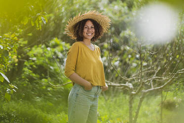 Smiling woman in straw hat standing by bare tree in vegetable garden - SBOF03148