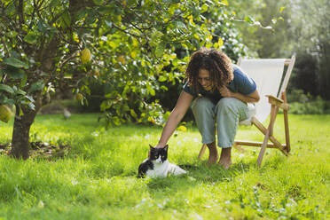 Lockenköpfige Frau streichelt Katze, während sie auf einem Stuhl im Garten sitzt - SBOF03164