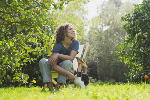 Lächelnde Frau mit lockigem Haar, die wegschaut, während sie auf einem Stuhl neben einer Katze im Garten sitzt - SBOF03166