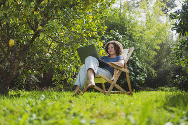 Lächelnde Frau schaut auf einen Laptop, während sie auf einem Stuhl im Permakultur-Garten sitzt - SBOF03168