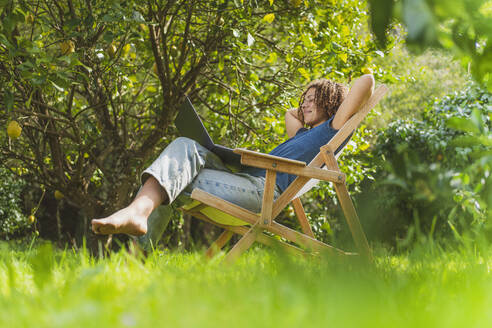 Lächelnde mittelgroße erwachsene Frau mit Händen hinter dem Rücken, die auf einen Laptop schaut, während sie auf einem Stuhl im Garten sitzt - SBOF03169