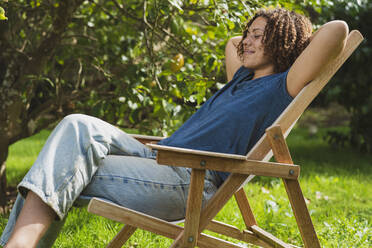 Lächelnde, lockig behaarte Frau mit geschlossenen Augen auf einem Liegestuhl im Garten ruhend - SBOF03170