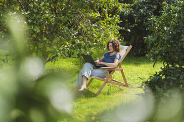 Mittlere erwachsene Frau, die einen Laptop benutzt und auf einem Stuhl inmitten von Bäumen im Garten sitzt - SBOF03172