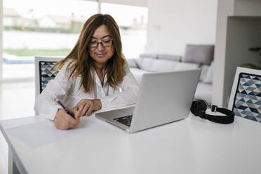 Berufstätige Frau, die mit Laptop und Kopfhörern zu Hause sitzt und auf Papier schreibt - EGAF02048