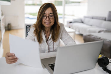 Unternehmerin hält Papier in der Hand, während sie zu Hause einen Laptop benutzt - EGAF02050