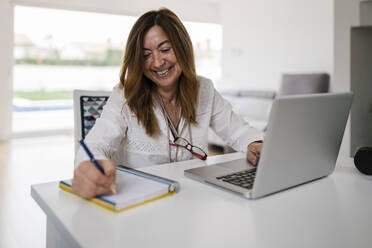 Lächelnde reife Frau, die in ihr Tagebuch schreibt, während sie einen Laptop zu Hause benutzt - EGAF02052