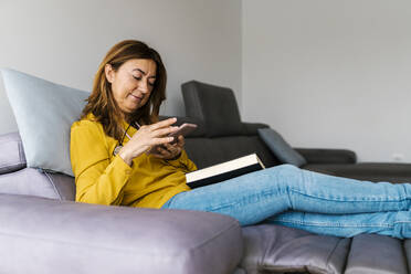 Reife Frau, die ein Smartphone mit einem Buch auf dem Schoß benutzt, während sie zu Hause auf einem Lehnstuhl sitzt - EGAF02057