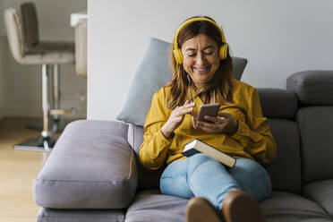 Smiling woman with headphones using smart phone while sitting on sofa in living room - EGAF02058