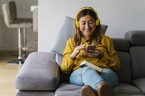Lächelnde Frau mit Kopfhörern, die ein Smartphone benutzt, während sie auf dem Sofa im Wohnzimmer sitzt - EGAF02058