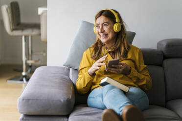 Lächelnde reife Frau, die wegschaut, während sie zu Hause über Kopfhörer Musik hört - EGAF02059