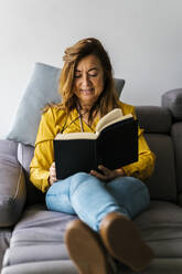 Woman reading book while sitting on sofa in living room at home - EGAF02062