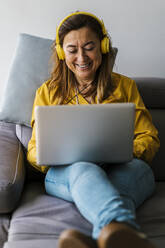 Smiling woman with headphones using laptop while sitting on sofa in living room - EGAF02063