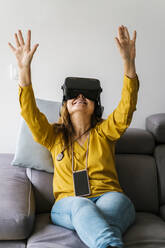 Reife Frau gestikuliert, während sie ein Virtual-Reality-Headset trägt und zu Hause auf dem Sofa sitzt - EGAF02066