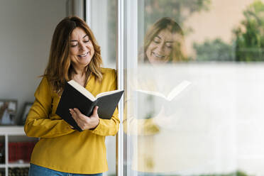 Lächelnde Frau, die ein Buch liest und sich zu Hause an ein Glasfenster lehnt - EGAF02067