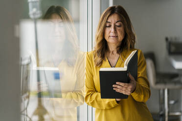 Frau liest ein Buch und lehnt sich an ein Glasfenster zu Hause - EGAF02068