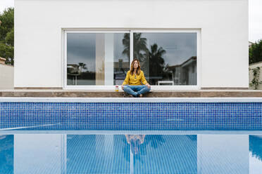 Mature woman meditating while sitting at poolside against house - EGAF02070