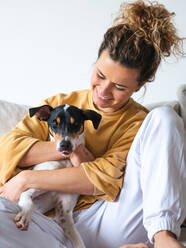 Fröhliche Frau mit lockigem Haar und legerer Kleidung sitzt mit dem gehorsamen Hund Ratonero Bodeguero Andaluz auf dem Sofa in einer hellen Wohnung - ADSF21209