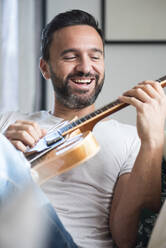 Glücklicher erwachsener ethnischer männlicher Musiker in Freizeitkleidung, der sich auf einem bequemen Sofa entspannt und zu Hause akustische Gitarre spielt - ADSF21238