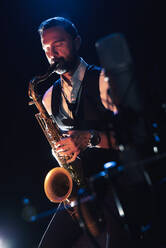 Seitenansicht eines konzentrierten bärtigen männlichen Musikers in elegantem Outfit, der Saxophon spielt, während er während eines Konzerts auf der Bühne steht - ADSF21240
