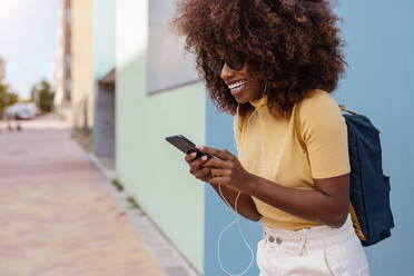 Black woman with afro hair listening to music on mobile in front of a blue wall - ADSF21247