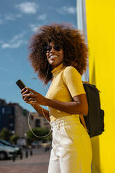 Schwarze Frau mit Afro-Haar, die vor einer gelben Wand mit dem Handy Musik hört - ADSF21248