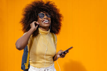 Schwarze Frau mit Afro-Haar, die vor einer orangefarbenen Wand mit dem Handy Musik hört - ADSF21250