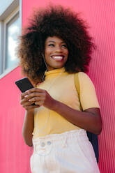 Black woman with afro hair listening to music on mobile in front of a pink wall - ADSF21252