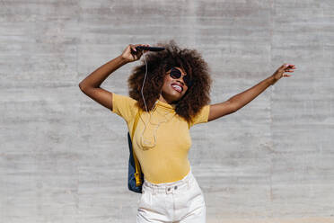 Schwarze Frau mit Afro-Haar, die vor einer grauen Wand mit dem Handy Musik hört - ADSF21257