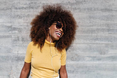 Schwarze Frau mit Afro-Haar, die vor einer grauen Wand mit dem Handy Musik hört - ADSF21258
