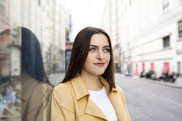 Smiling beautiful woman looking away while standing by glass wall in city - JCCMF01396