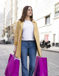 Young woman carrying magenta colored shopping bags while walking on road in city - JCCMF01397